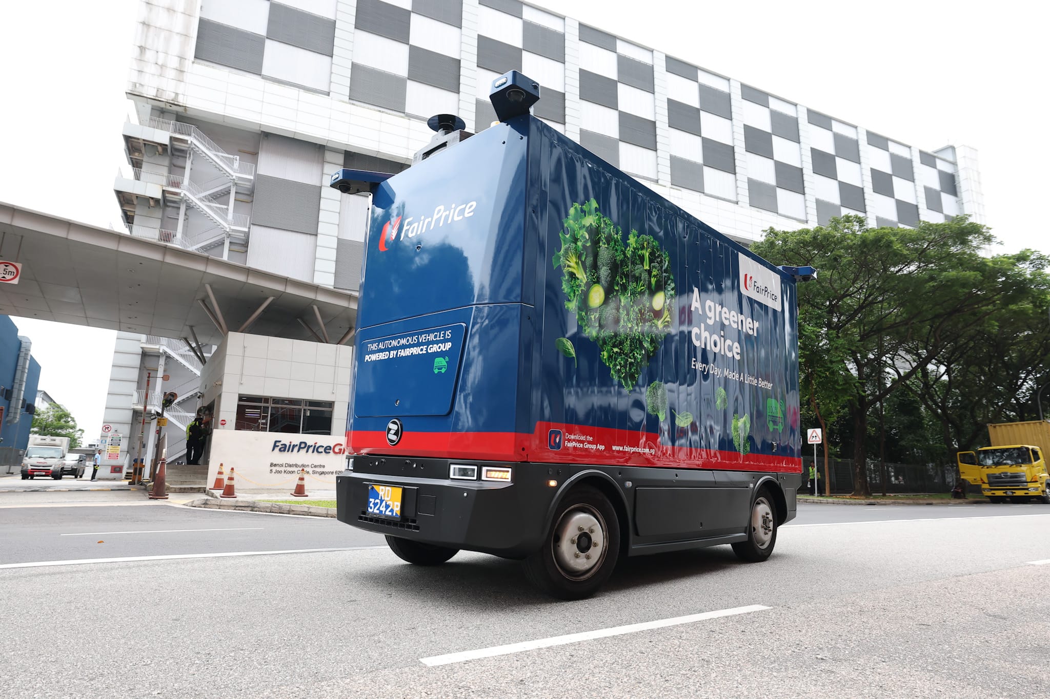 A blue FairPrice autonomous delivery vehicle with sensors on the roof drives on a road past an industrial building.